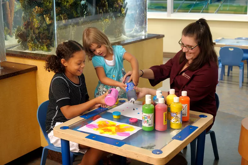 Two children and an adult woman sitting at a low arts and crafts table doing crafts.