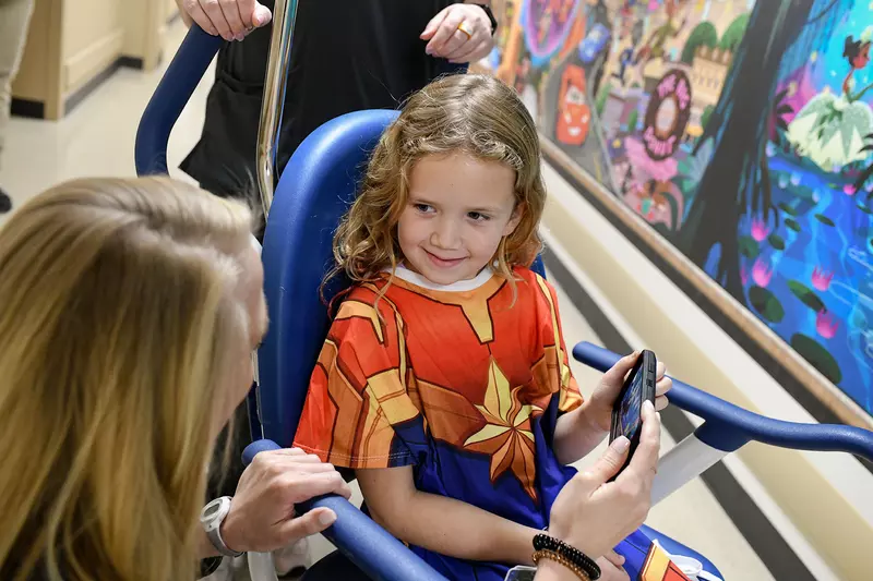 Little girl in a wheelchair at AdventHealth for Children wearing a Captain Marvel gown and using the Disney Team of Heroes mobile app.