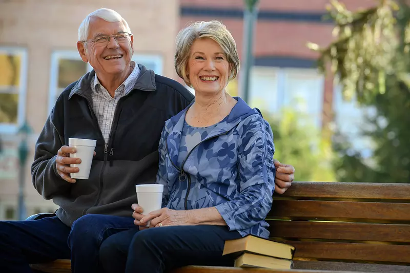 Elderly couple sitting on a park bench drinking coffee.