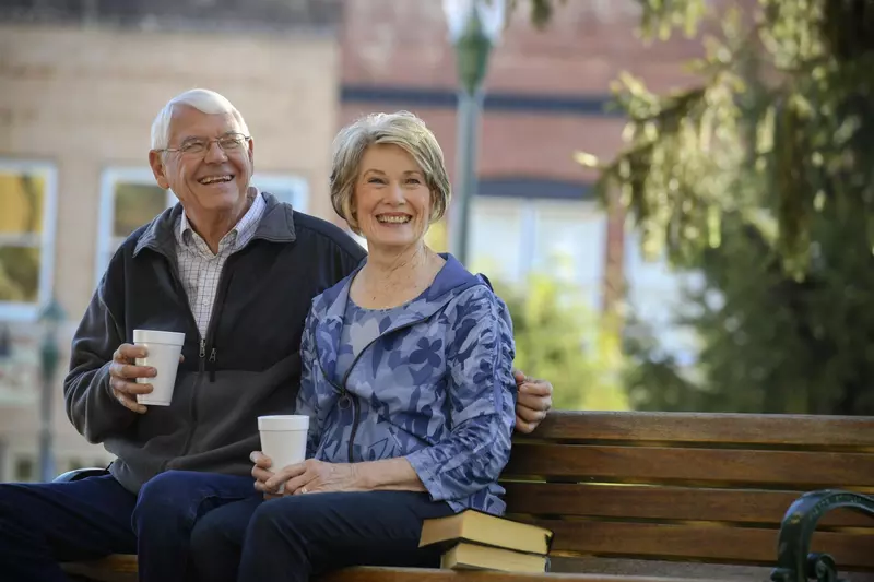 Elderly couple sitting on a park bench drinking coffee.