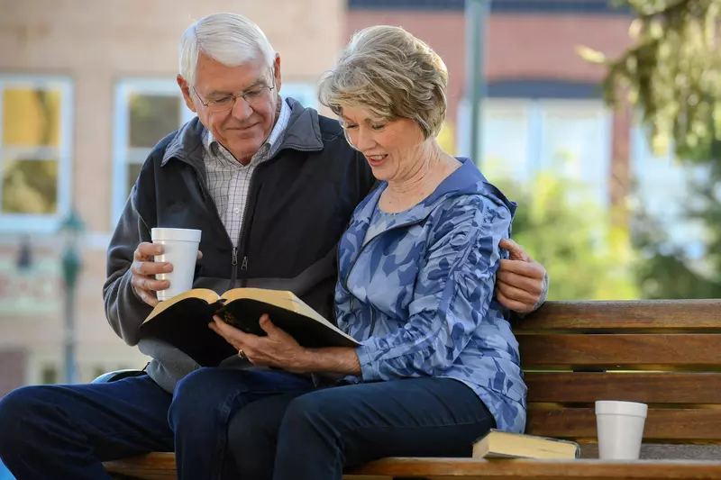 Elderly Couple sitting on a park bench reading the Bible.