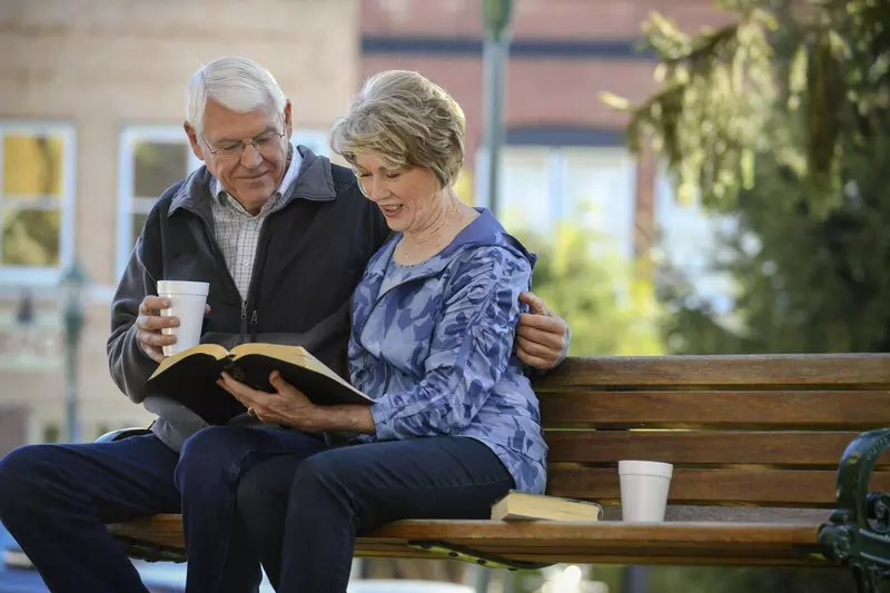 Elderly couple sitting on a park bench reading the bible. 