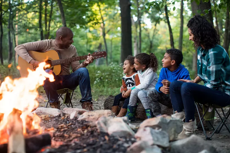 A family singing along while the father plays the guitar around the fireplace in the forest.