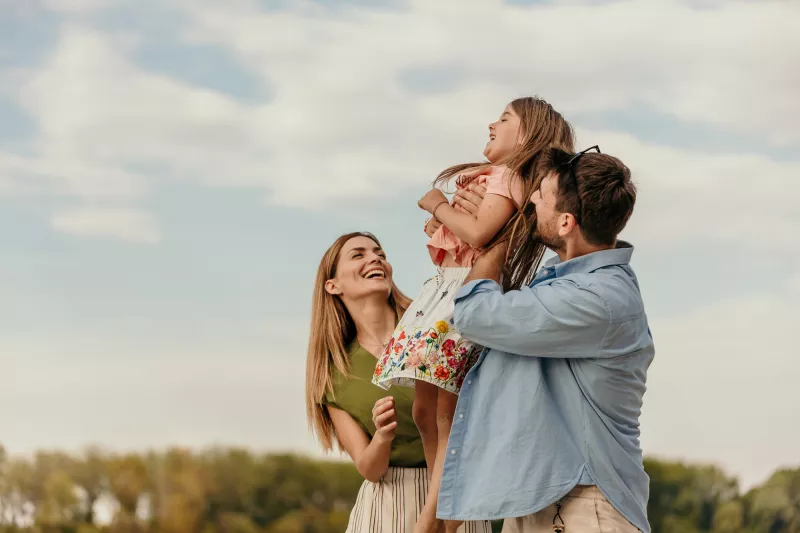 A family of a mother, father and daughter smiling while outdoors.