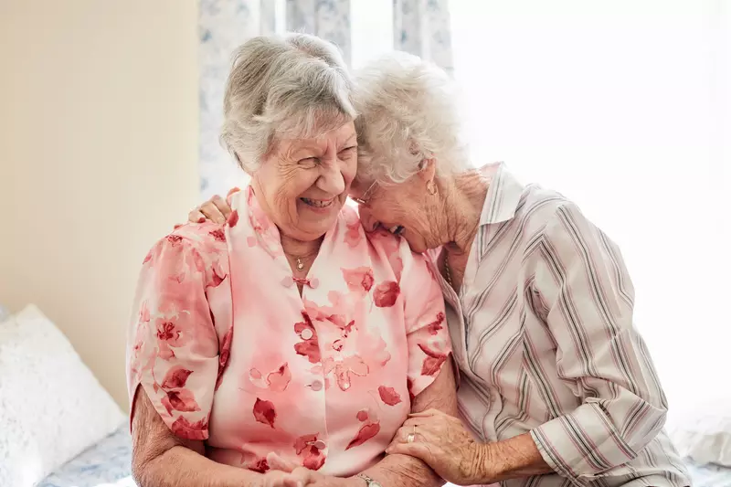 Two older women laugh together while sitting on a bed.