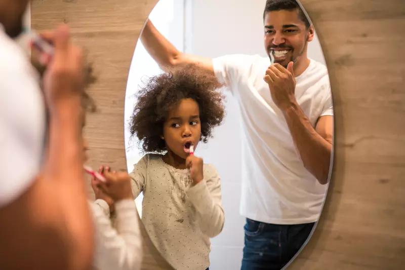A father and daughter brush their teeth looking in a mirror.