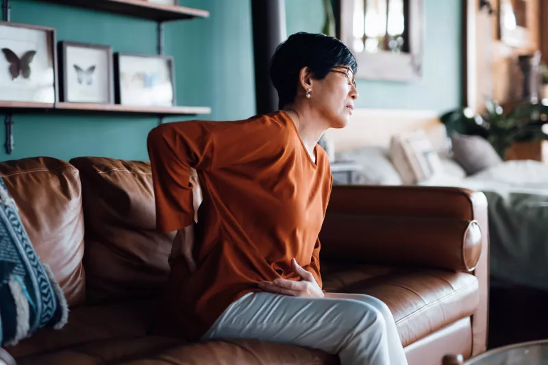 A woman holding her back in pain while sitting on a sofa.