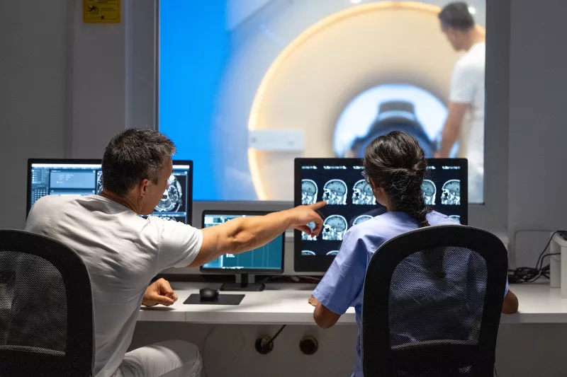 A male and female imaging technician looking at CT scans of a patient's head in the foreground while another male technician is prepping the patient in the CT machine in the background.
