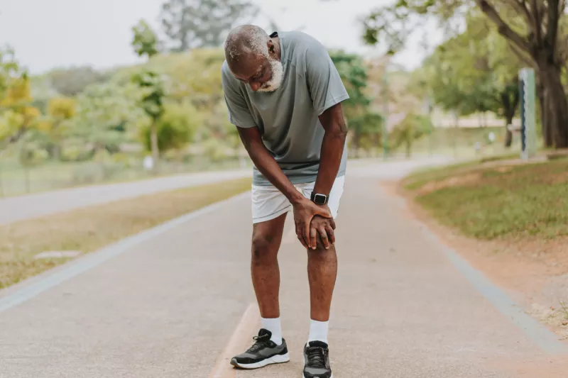 A senior black man takes a break from exercising outside and places his hands on his left knee.