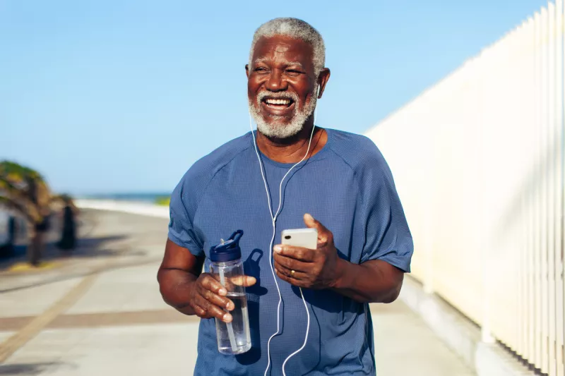 An older black man using headphones plugged into his phone while walking outdoors and holding a water bottle.