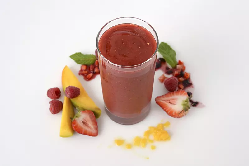a tall glass of smoothie, surrounded by chopped mango and mixed berries