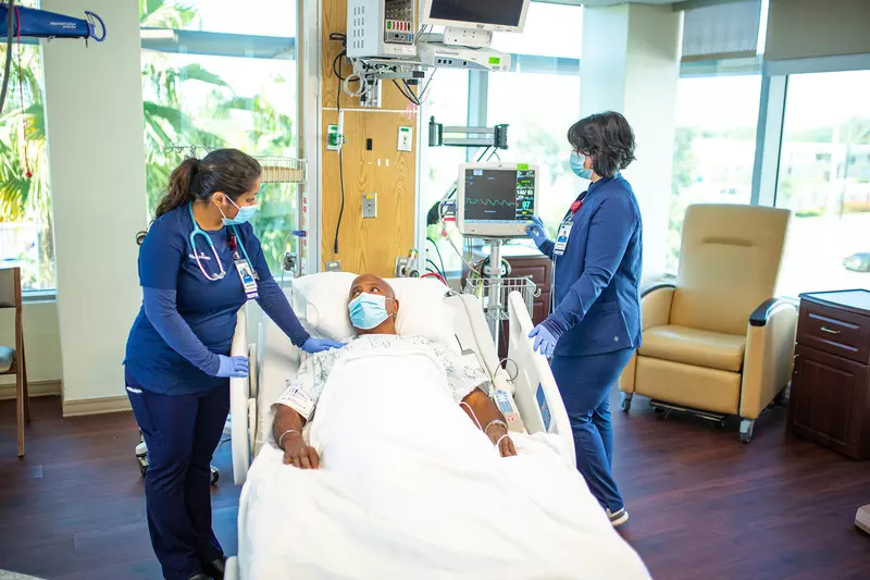 Man in the ICU talking to a nurse while another nurse checks a monitor. All are wearing masks.