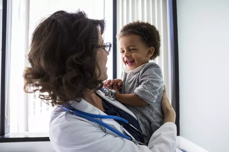 A female pediatrician holds a smiling infant in an exam room. 