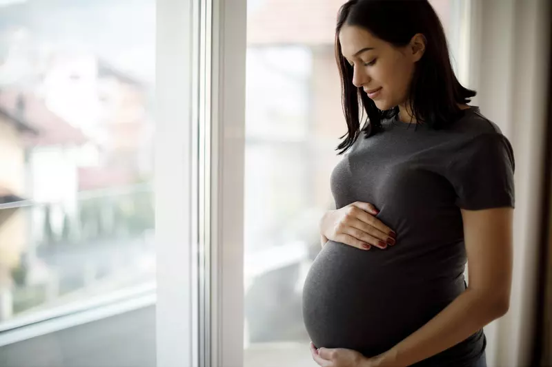 A pregnant young woman holds her belly standing near a window