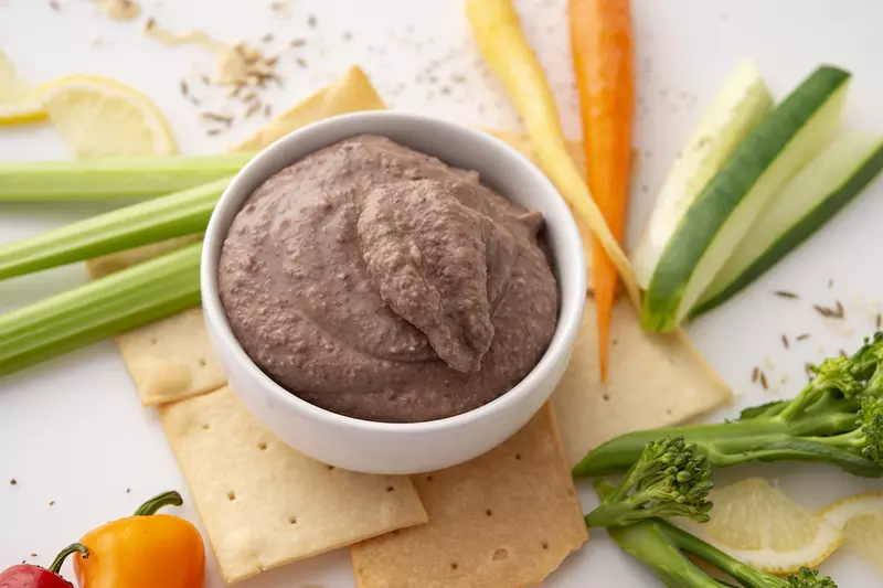 White bowl of black bean hummus surrounded by vegetable sticks and crackers.