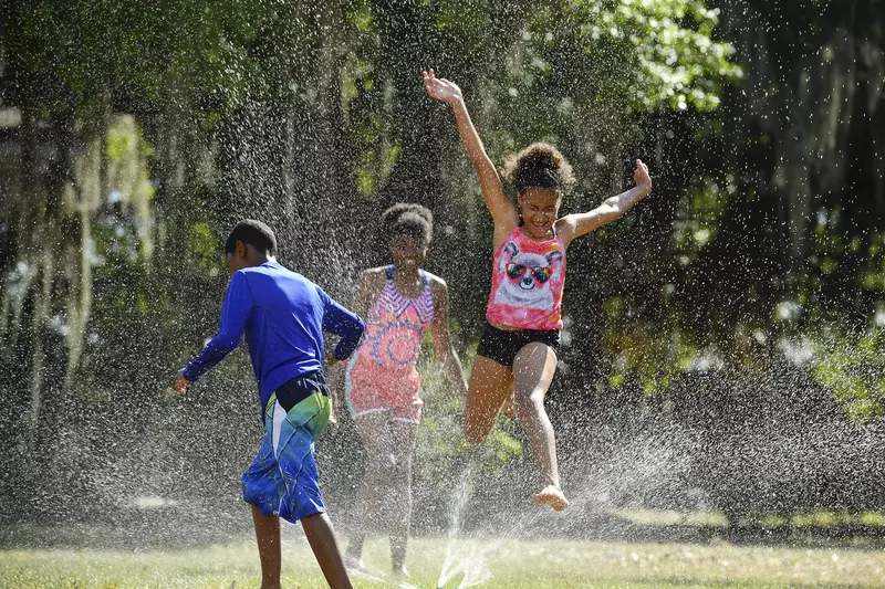 Young African American children run and jump through an outdoor sprinkler.