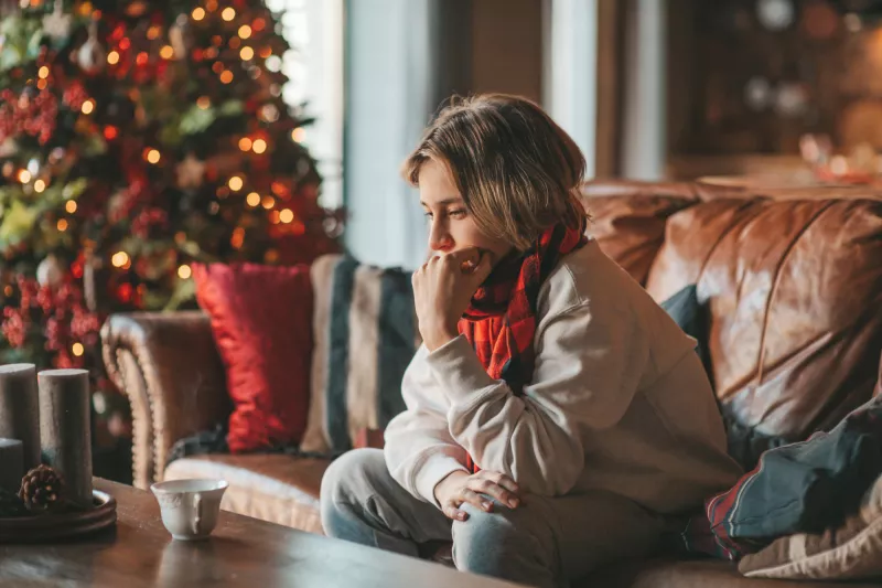 A teen boy sits on a couch at home looking introspectively near a Christmas tree.