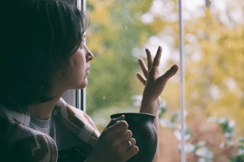 Woman looking out and touching window while indoors while wrapped in a blanket and holding a mug.