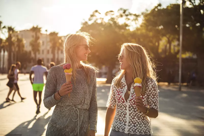 Women smiling at each other while holding ice cream cones and walking outdoors.