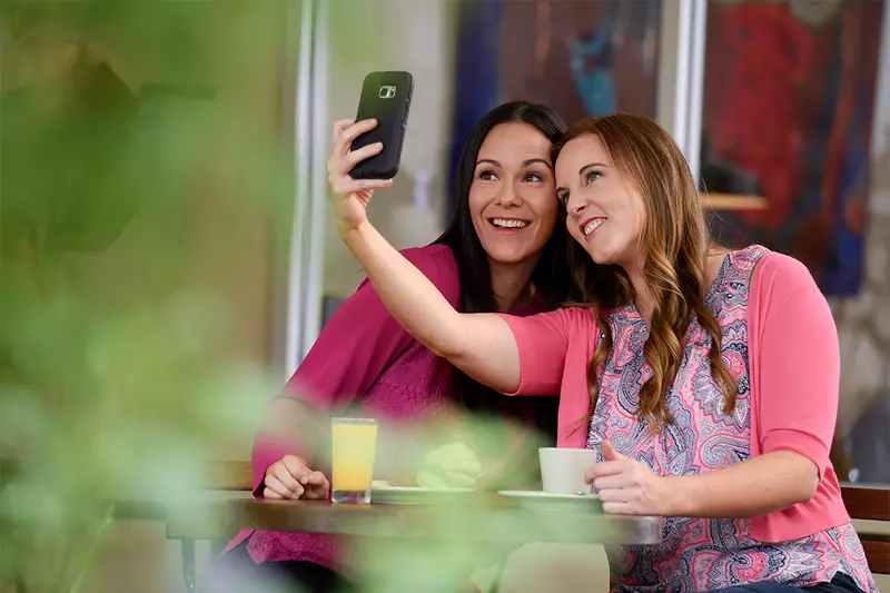 Women sitting at a table taking a selfie photo with a camera phone.