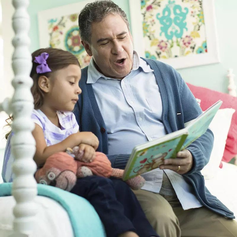 A grandfather reads a children's book to his granddaughter in her bedroom.