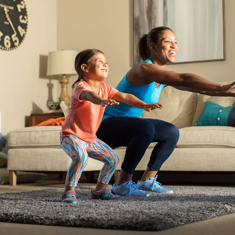 A mother and her young daughter exercise together in the living room.