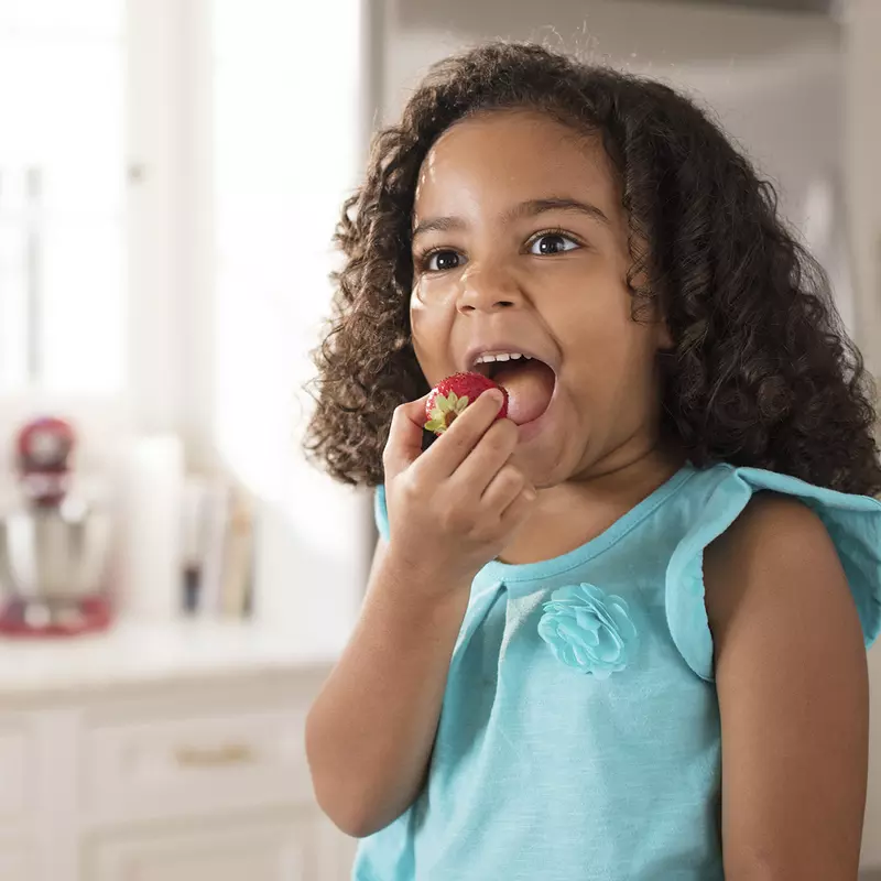 A young African American girl eats strawberries in the kitchen.