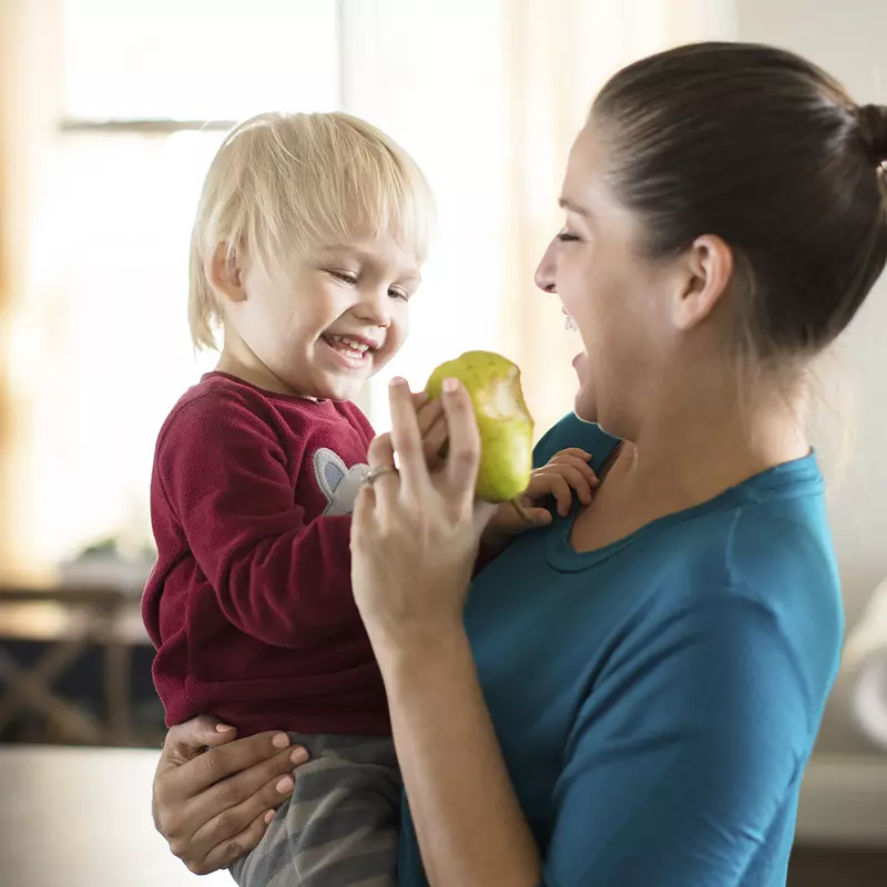A Caucasian mom feeds her son a pear in the kitchen.