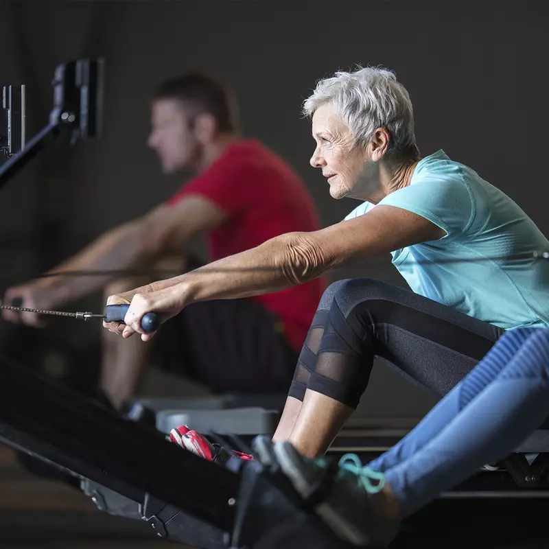 An elderly Caucasian woman exercises on a rowing machine at the gym.