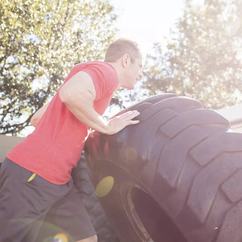 A young Caucasian man exercises outdoors by flipping large tires.