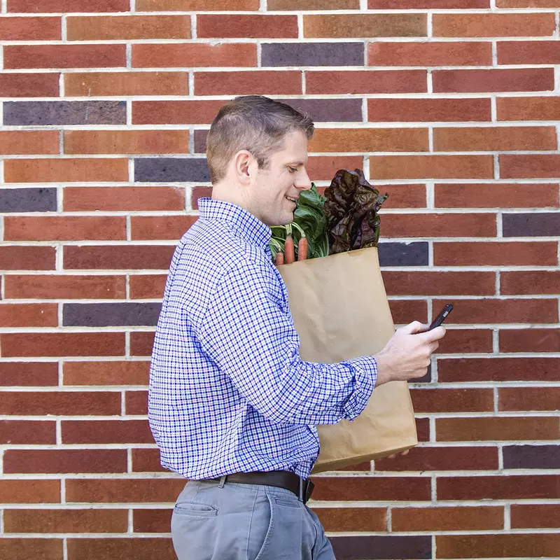A young Caucasian man checks his cell phone while carrying a bag of fresh produce.
