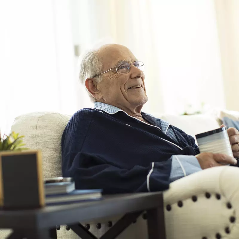 An elderly Caucasian man sits on the sofa with a mug of coffee