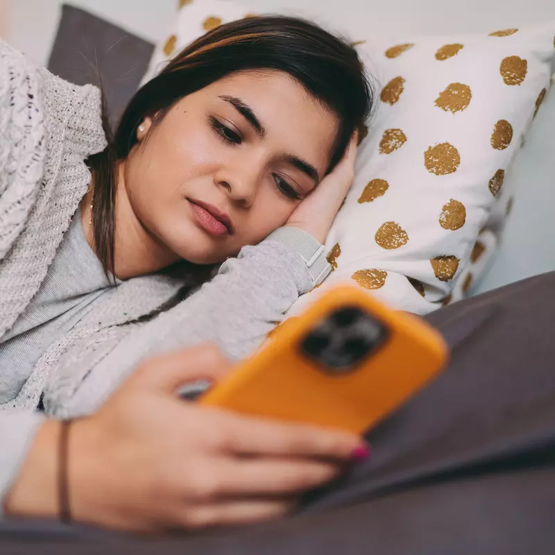 A Woman Lays on a Bed Looking at Her Cell Phone.