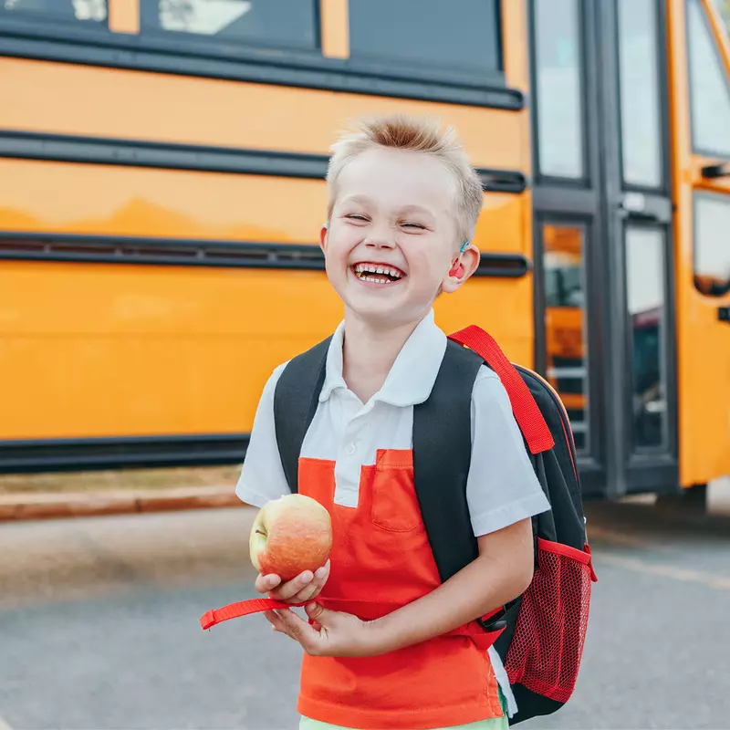 A Small Boy Laughs as He Gets Off a School Bus with an Apple in Hand.