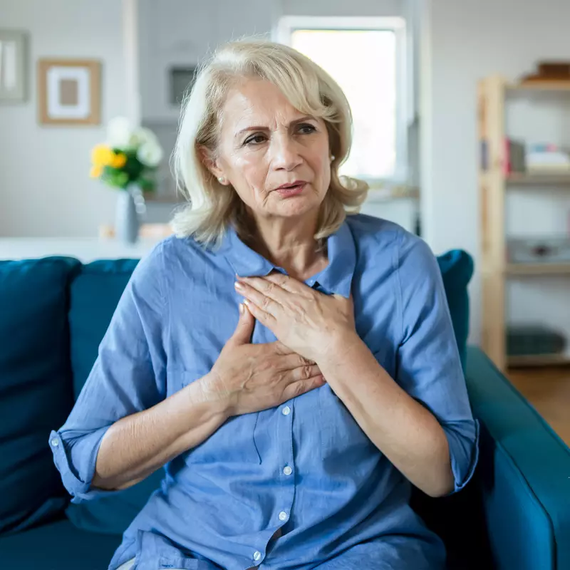 A Woman Sits in Her Living Room With Her Hands on Her Chest, Feeling Her Heartbeat.