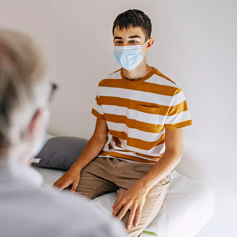A Teenager Wearing a Face Mask Speaks to His Physician.