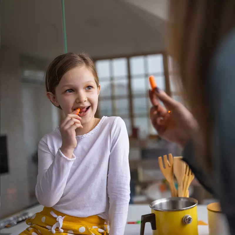A Mother Hands Her Daughter Carrot Sticks to Eat in the Kitchen.