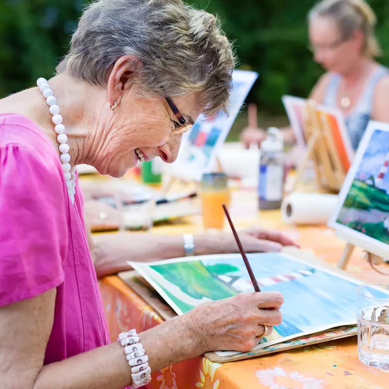 A Grandmother Paints a Watercolor Painting Based on an Example on a Tablet.