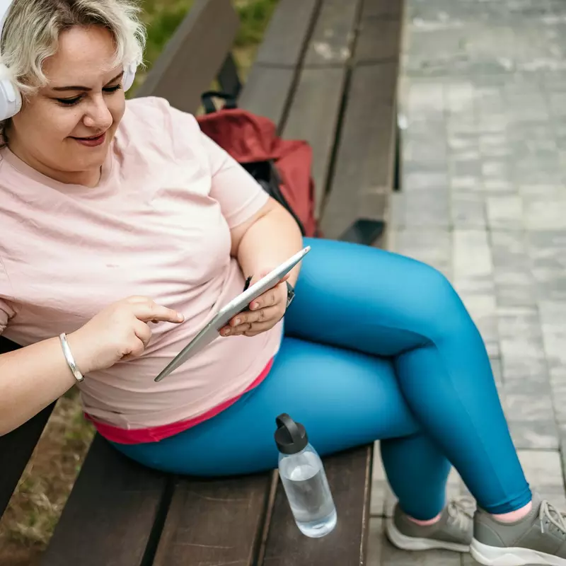 A Woman Sits on a Park Bench Scrolling Through Her Tablet Listening to Music