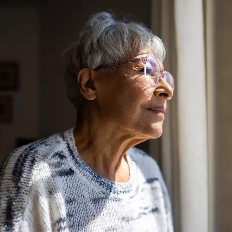 A Senior Woman Looks Out The Window of Her Home with a Smile