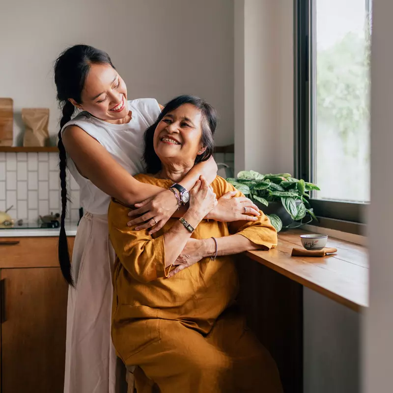 A Mother and Adult Daughter Embrace in the Kitchen in Front of a Window