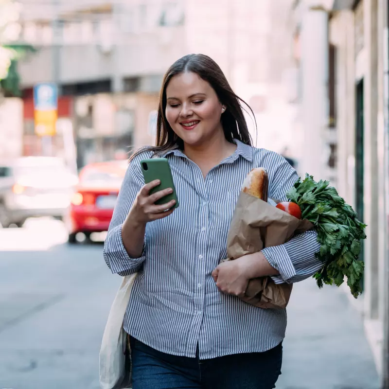 A Woman Walks Down the Sidewalk Checking Her Phone While Carrying Groceries