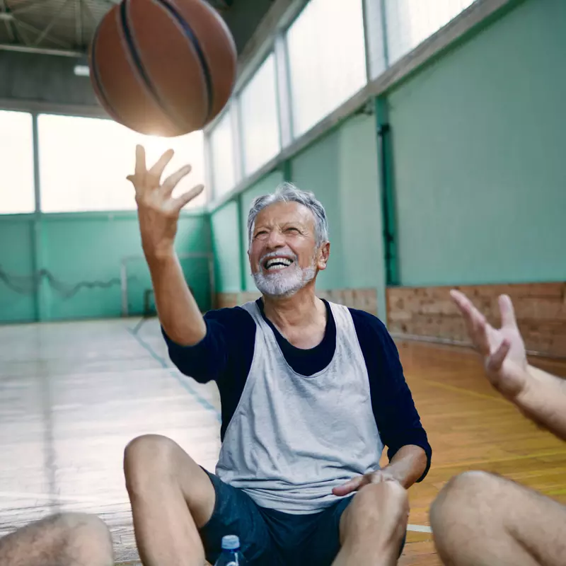 A Trio of Senior Men Sit on a Gymnasium Floor While One of Them Tosses a Basketball in the Air
