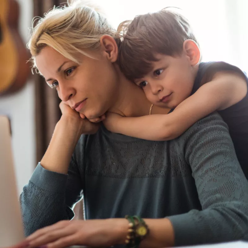 A Mom Stares at A Computer Screen While Her Young Son Hugs Her From Behind