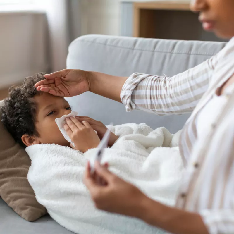 A Small Child Blows His Nose Laying on a Couch While His Mother Checks His Temperature