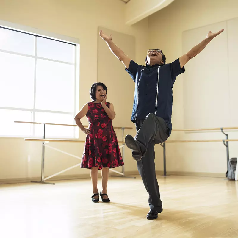 A Hispanic man leaps for joy in a dance studio while his wife smiles on.