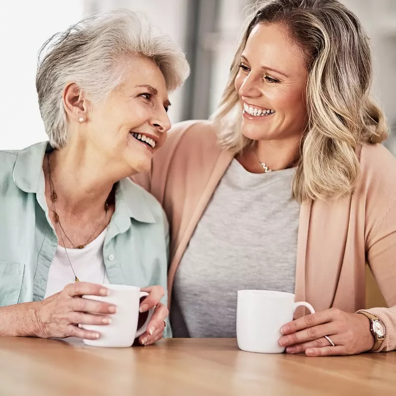 Adult woman with her arm around her mother, both smiling and holding white mugs.