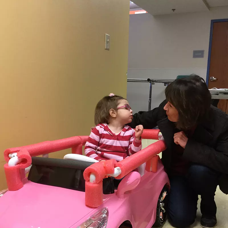 A little girl simulating to drive a pink car toy while kissing her mother.
