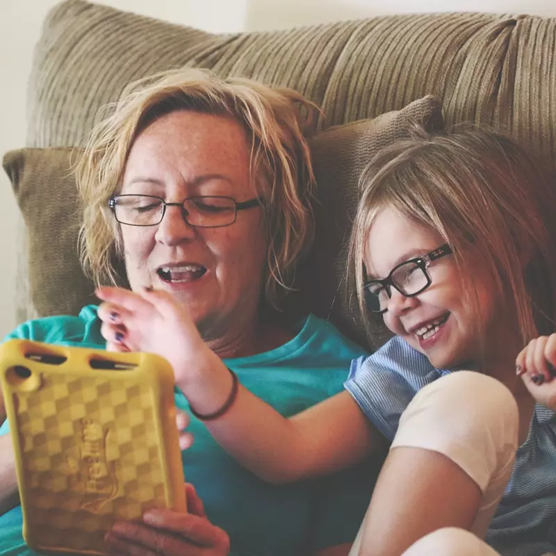 A grandmother playing with her grand-daughter in the couch with a tablet. 