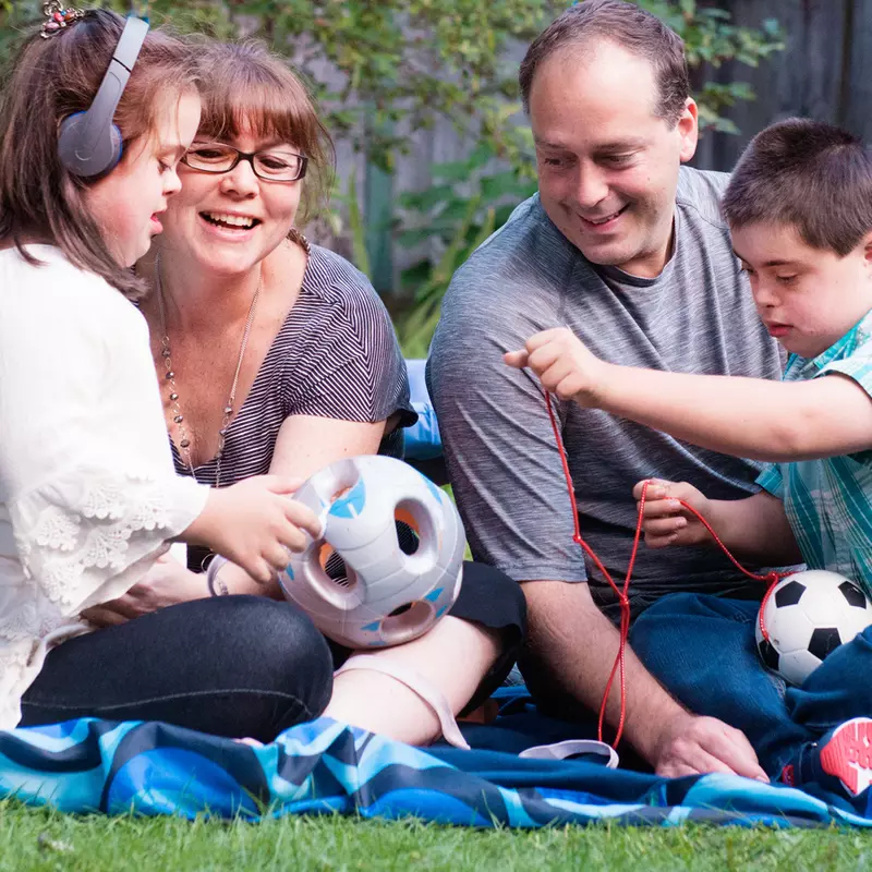 A family of four sitting on a blanket outside, playing with a soccer ball.